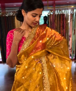 A Mustard yellow dual tone tissue silk saree with a sublte sheen , featuring a contrasting pale gold border highloghted with intricate gota work in geometric and froral motifs drape elegantly over a shoulder with soft pleats
