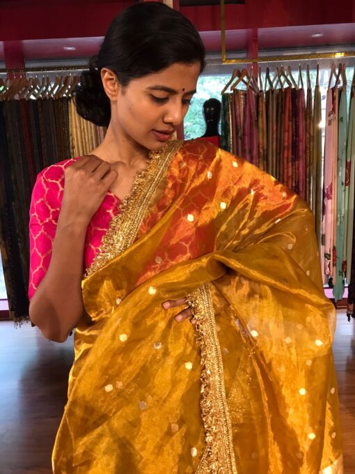 A Mustard yellow dual tone tissue silk saree with a sublte sheen , featuring a contrasting pale gold border highloghted with intricate gota work in geometric and froral motifs drape elegantly over a shoulder with soft pleats